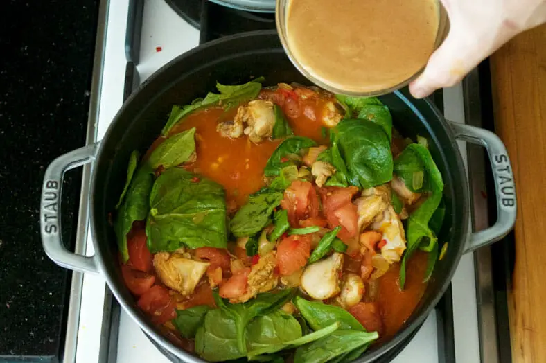 The next step to making Muamba Nsusu, a Congolese Peanut & Palm Oil Stew, is once your minced red chilies, cumin powder, tomatoes and spinach have had some time to cook together (and your spinach has started to soften), add in your peanut butter and mix everything around to distribute it well.